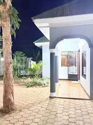 a white house with a porch and a tree at Cozy-Homey with solar system -Firepit- Free Parking-Gardenswing in Arusha
