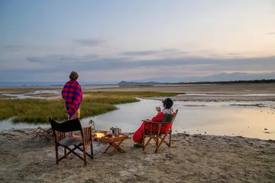 two people sitting in chairs on a beach with a table at Kisima Ngeda Camp in Mangola